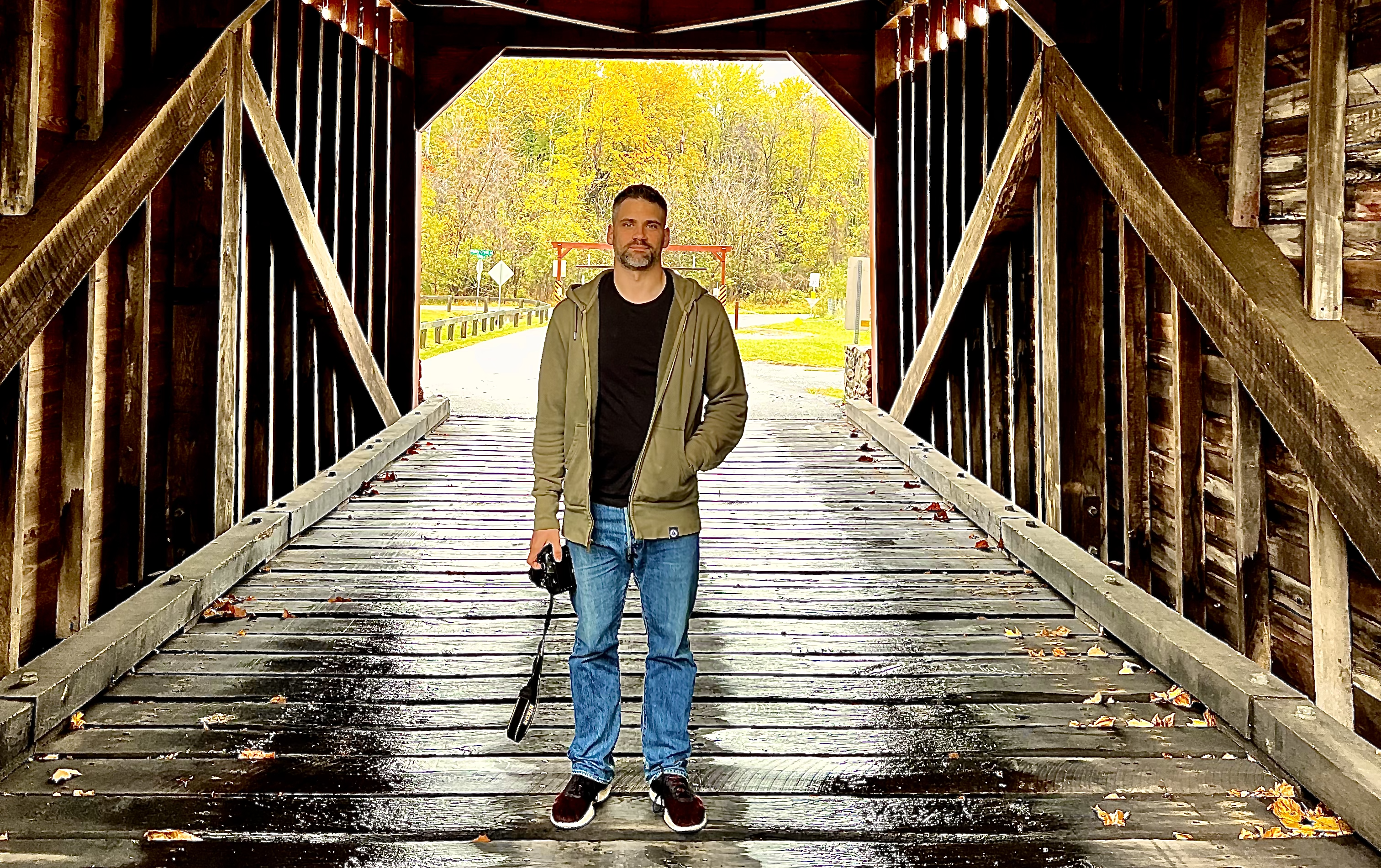 A man standing on a wet covered bridge holding a camera in his left hand